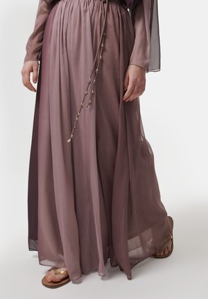 Langer, fließender Rock in einem Farbverlauf sanfter Mauve-Töne, aus transparentem Stoff mit einem elastischen Bund und dekorativen Bindungen. Sandalen sind sichtbar.
