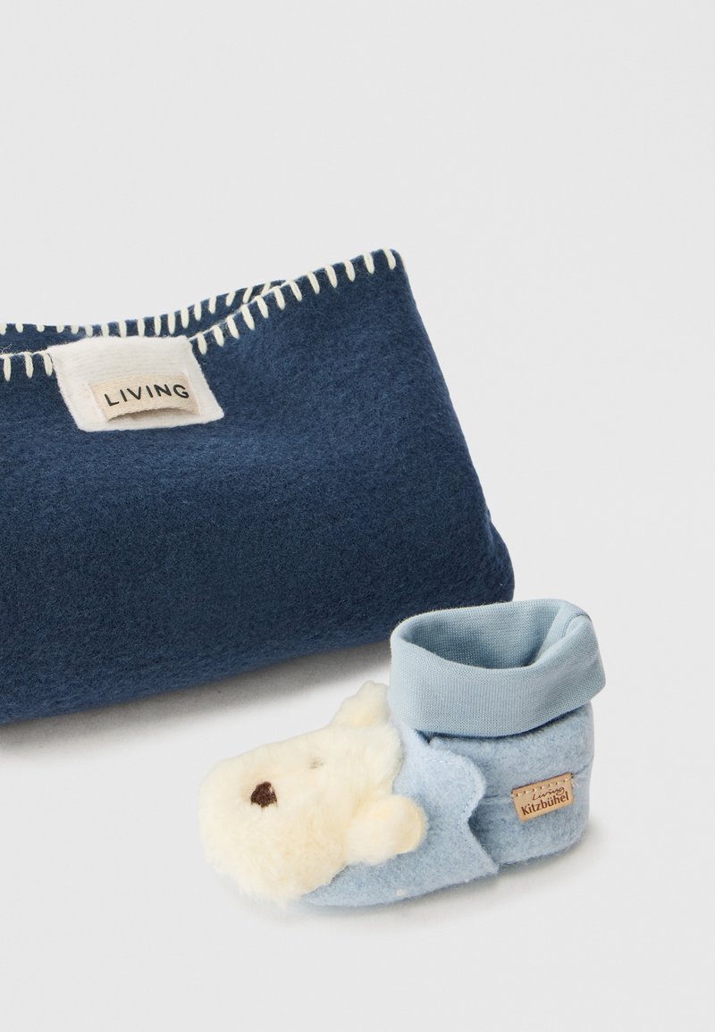 Μπλε παντόφλα παιδιών από μαλακό τσόχας, με σχέδιο από αρκουδάκι και αφράτες λεπτομέρειες. Σκούρα μπλε φλις τσάντα με άσπρα ράμματα και ετικέτα.