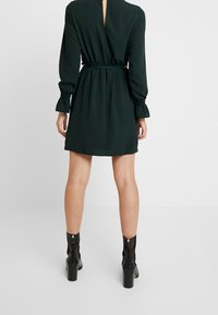 Grön långärmad klänning med åtdraget midja, med en nyckelhålsdetalj i ryggen och elastiska manschetter. Kombinerad med svarta klackiga ankelstövlar.