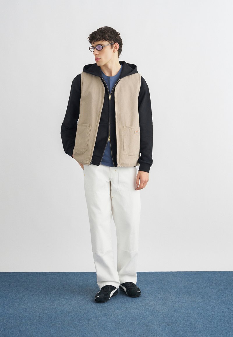 Joven con gafas, sudadera negra con capucha, chaleco beige, pantalones blancos y zapatillas negras, de pie sobre una alfombra azul con las manos en los bolsillos.