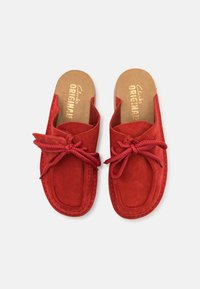 Red suede slip-on shoes with a rounded toe, featuring a bow detail and beige cork insole with embossed brand name. Stitched accents around edges.