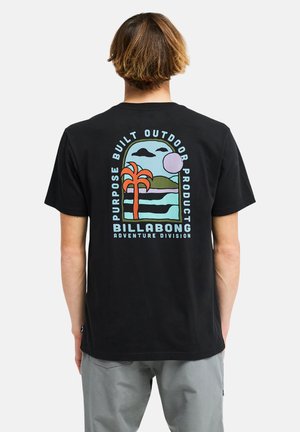 Homme aux cheveux bruns courts portant un t-shirt noir avec un motif coloré de palmier et d'océan et le texte Billabong au dos.