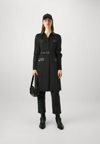 Zwarte trenchcoat met knoopzakken, een ceintuur die om de taille is cinched, en zilveren hardware-accenten. Gecombineerd met zwarte jeans, enkellaarsjes en een pet.