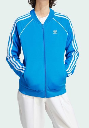 Osoba ubrana w jasnoniebieską kurtkę Adidas na zamek błyskawiczny z białymi paskami na rękawach oraz dopasowane białe spodnie, ręce w kieszeniach kurtki.