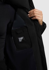 Käsi hoiab avatud musta tepitud jope, näidates sisemist taskut, millel on Khujio™ logo täkk mustal voodil.