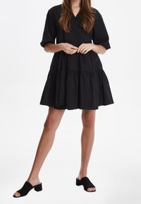 Femme portant une robe mini noire à volants avec des manches bouffantes et des mules à talons blocs noirs à bout ouvert, se tenant les mains croisées à la taille.