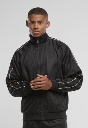 Ung mand med kort hår iført en sort lynlåstrøje med hvide striber, stående med hænderne foldet foran mod grå baggrund.