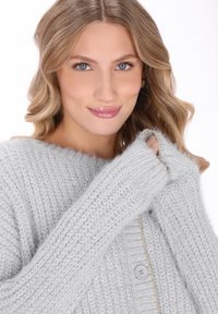 Młoda kobieta z falującymi blond włosami, nosząca przytulny, jasnoszary, dziergany sweter, delikatnie trzymająca rękaw blisko twarzy, uśmiechająca się łagodnie.