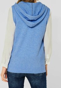 Lichtblauwe gebreide mouwloze hoodie vest met ribbelzoom, met een zachte textuur en een capuchon aan de achterkant over een witte lange mouwen shirt.