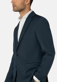 Mann in einem dunkelblauen Blazer und einem weißen Hemd, mit Händen in den Taschen, aus der Brustansicht vor einem einfarbigen hellen Hintergrund zu sehen.