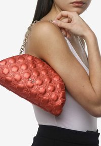 Un sac à main matelassé de couleur rouge rosé avec une texture brillante, orné de coutures à motif floral et une bandoulière en chaîne dorée, porté sous le bras.