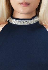 Närbild på en kvinna som bär en marinblå halterneckklänning med hög krage prydd med silver- och pärlblommor som dekoration.