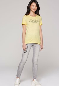 Gelbes kurzärmliges T-Shirt mit schwarzem Text, kombiniert mit hellgrauen Skinny Jeans mit seitlichem Text und einem used Look. Weiße Sneakers.