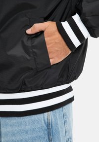 Čierna bomber bunda s hladkou textúrou, s bielymi a čiernymi pásikmi na rebrovaných manžetách a detailom na vrecku, nosená cez svetlomodré džínsy.