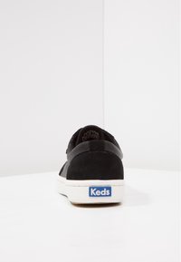 Černé semišové tenisky s bílou podešví a zadním štítkem, vyznačující se modrým logem "Keds". Hladká textura a nízký profil.
