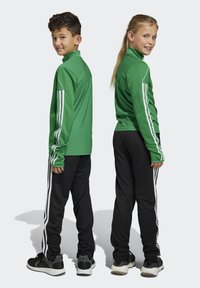 Grüne sportliche Reißverschlussjacken mit langen Ärmeln, schwarze Hosen und weiße Seitenstreifen; getragen von zwei Kindern, die von hinten und von der Seite schauen.