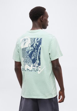 Homme de dos portant un t-shirt vert clair avec un motif abstrait bleu et blanc et un texte dans le dos, associé à un short foncé.