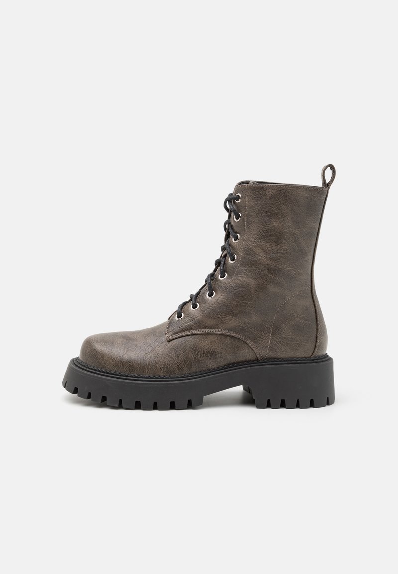 Ruskea tekonahkasaapas nilkkamallia, jossa on mustat nauhat, pyöreä kärki, paksu musta pohja ja vedettävä lenkki takana. Rakeinen pinta, jossa on hieno kiilto.