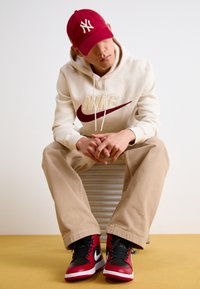 Cappellino rosso con logo NY, felpa beige con marchio Nike, pantaloni beige e sneakers rosso/nero. Seduto su una superficie a righe, sfondo neutro.