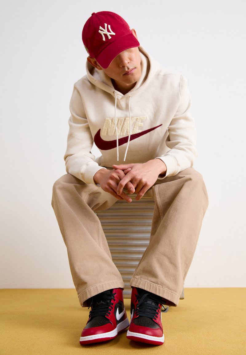 Rote Kappe mit NY-Logo, beiges Hoodie mit Nike-Branding, beige Hose und rot/schwarze Sneaker. Sitzt auf einer gestreiften Oberfläche, neutraler Hintergrund.