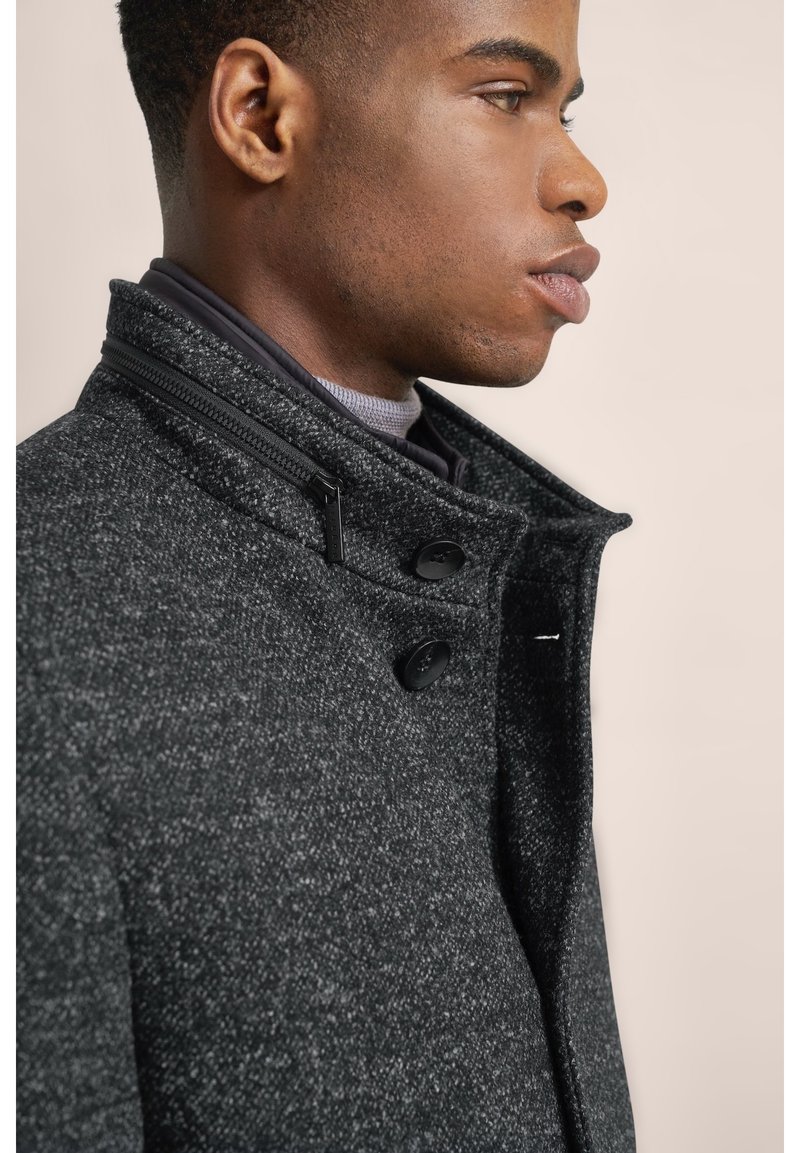 Cappotto grigio texturizzato con collo alto, zip e due bottoni neri. Presenta un design aderente, esposto su uno sfondo neutro.