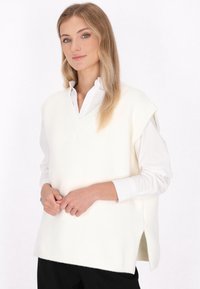 Gilet bianco in misto lana con scollatura a V, maniche corte e spacchi laterali, indossato sopra una camicia bianca con bottoni. Finitura morbida e testurizzata.