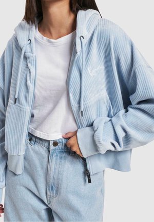 Osoba nosząca jasnoniebieską prążkowaną bluzę z kapturem na suwak, na białym krótkim t-shircie i jasnoniebieskich jeansach z wysokim stanem, z jedną ręką w kieszeni.
