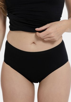 Frau trägt schwarze Baumwollunterwäsche mit hohem Bund und ein schwarzes Oberteil, hebt das Oberteil leicht an, um Taille und Bauch zu zeigen.