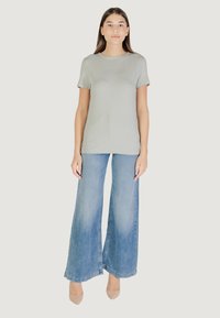 Maglietta aderente grigia chiara con logo "GUESS" in rilievo, abbinata a jeans blu a gamba larga e tacchi nudi, su uno sfondo neutro.