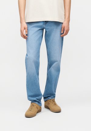 Muž oblečený v svetlomodrých džínsach s rovnými nohavicami a béžových teniskách, stojaci pred jednoduchým bielym pozadím.