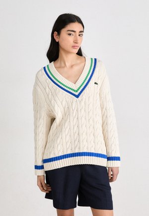 Crème kabelgebreide trui met een V-hals, voorzien van groene en blauwe gestreepte accenten op de kraag en zoom. Gepaard met donkere shorts.