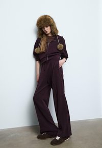 Burgunderroter Reißverschluss-Hoodie mit großen, runden Fellpompons, kombiniert mit einer weit geschnittenen Hose. Dazu eine pelzige braune Mütze und dunkle, schnürbare Schuhe.