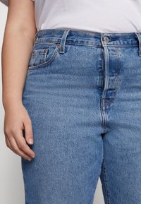 Jeans en denim bleu taille haute avec une finition délavée, présentant des rivets en cuivre, un design à cinq poches et des passants de ceinture. Tissu texturé visible.