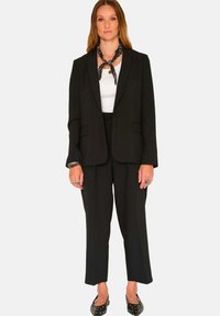Blazer noir ajusté avec une fermeture à un bouton, pantalon assorti et top blanc ; comprend une écharpe noire à motif et des chaussures à pois.