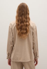 Beige corduroy overhemd met lange mouwen, gestructureerde stof, zijsplitten en knoopmanchetten. Weergegeven van achteren, met golvend haar zichtbaar.
