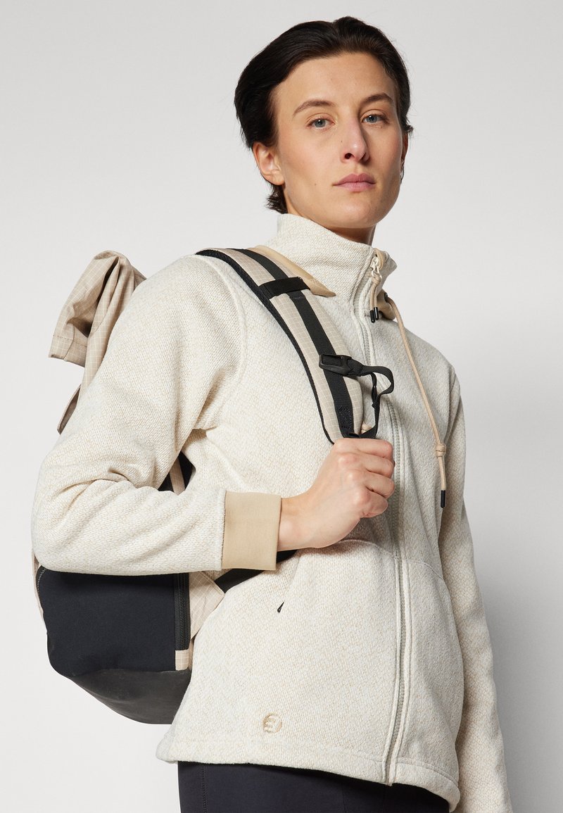 Veste en molleton beige clair zippée avec un motif en chevrons texturé, des poignets chauds, et un sac à dos noir et beige avec des bretelles réglables.