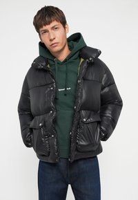 Chaleco de plumas negro con acabado brillante, botones de presión y dos bolsillos frontales, superpuesto a una sudadera verde con texto blanco. Pantalones de mezclilla informales.