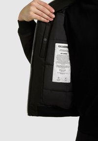 Schwarze isolierte Jacke mit glattem Außenstoff, gesteppter Innenfutter, silbernem Reißverschluss und Pflegeetikett, das auf der Innenseite sichtbar ist.