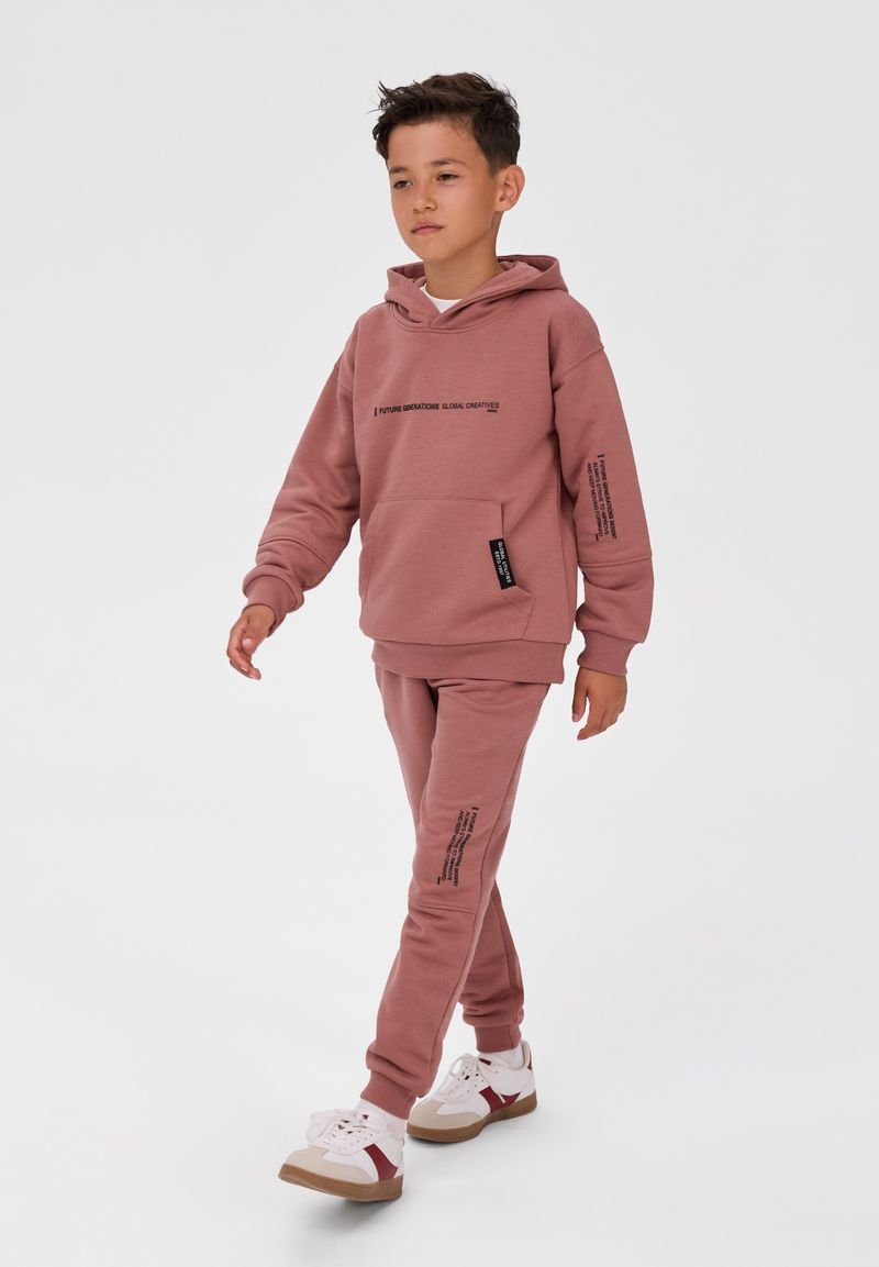 Garçon marchant portant un sweat à capuche et un pantalon rose assortis avec un texte noir et des baskets blanches avec des détails rouges et beige.