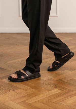 Persona con pantaloni neri e sandali neri con cinturini incrociati che cammina su un pavimento in parquet di legno marrone chiaro all'interno.