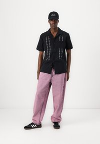 Svart kortermet skjorte med hvite vertikale mønstre, kombinert med rosa baggy jeans og svarte sneakers med hvite striper. Svart caps er også brukt.