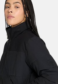 Giacca a vento nera con colletto alto, texture trapuntata e chiusura con zip laterale. Presenta un logo ricamato nero sulla manica.