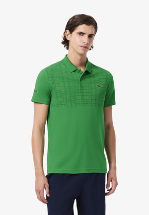 Jeune homme portant un polo vert avec un motif géométrique sur la partie supérieure de la poitrine et un pantalon bleu foncé, se tenant contre un fond blanc.