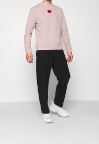 Sudadera de color rosa claro con un pequeño parche de logo rojo, combinada con pantalones negros de corte holgado y zapatillas deportivas blancas.