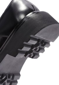 Schwarze Lederstiefel mit einer dicken Gummisohle, die ein tiefes Profilmuster aufweist. Das Obermaterial hat eine glatte Oberfläche und eine abgerundete Zehenform.
