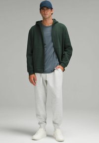 Donkergroen zip-up hoodie over een grijs t-shirt, gecombineerd met lichtgrijze joggingsbroek en witte sneakers, met een marineblauw pet. Eenvoudig en casual design.
