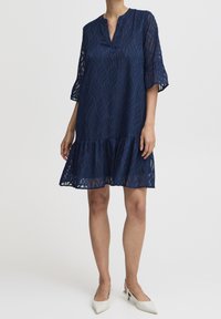 Laivansininen mekko, jossa on pitkät, puolisiistot hihat ja röyhelöhelma; siinä on teksturoitu lehtikuvio ja pyöreä kaula-aukko.