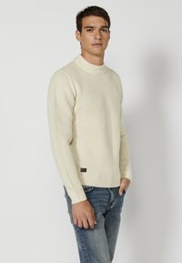 Crème-kleurige gebreide trui met een textuurpatroon, hoge kraag en ribbelmanchetten. Heeft een klein bruin leren label op de zoom.