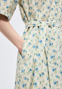 Χέρι ατόμου στην τσέπη, που φοράει κρεμ φόρεμα με μπλε λουλουδένιο σχέδιο, κοντά μανίκια, κουμπιά μπροστά και υφασμάτινη ζώνη δεμένη στη μέση.