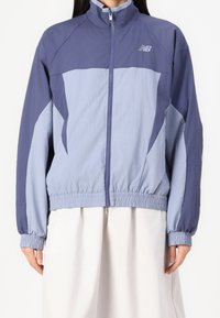 Žena oblečená v dvoubarevném svetle a tmavě modrém bomberu se zipem, s elastickými manžetami a lemem, spárovaná s bílou sukní.